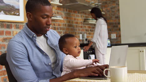 Father-And-Baby-Daughter-Use-Laptop-As-Mother-Prepares-Meal