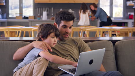 Father-And-Son-Sitting-On-Lounge-Sofa-Using-Laptop-Together