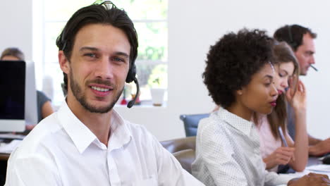 Man-wearing-headset-in-an-open-plan-office-smiles-to-camera