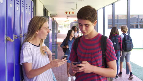 Two-teenage-friends-talking-use-phones-in-school-corridor