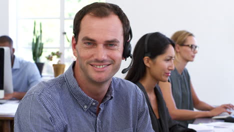 White-man-in-headset-smiles-to-camera-in-open-plan-office
