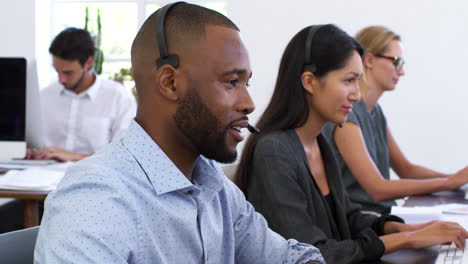 Black-man-in-headset-smiles-to-camera-in-open-plan-office
