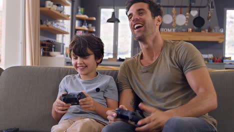 Father-And-Son-Sitting-On-Sofa-In-Lounge-Playing-Video-Game