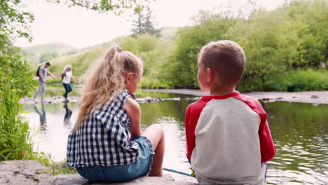 Zeitlupenaufnahme-Von-Kindern,-Die-Auf-Felsen-Sitzen-Und-Mit-Netzen-Im-Fluss-Im-Britischen-Seengebiet-Fischen