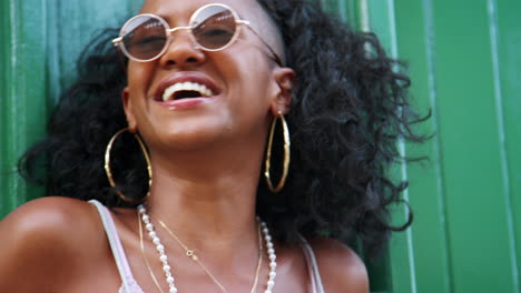 Young-black-woman-wearing-sunglasses-leaning-on-green-wall-in-the-laughing,-head-and-shoulders