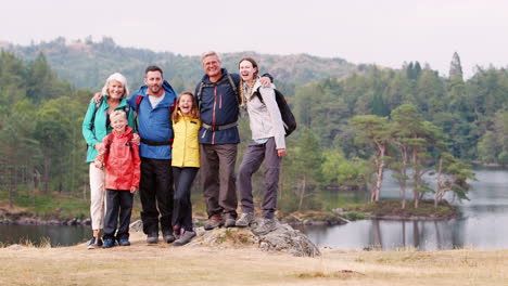 Multi-generation-family-standing-on-rocks-by-a-lake-in-the-countryside-looking-to-camera,-Lake-District,-UK