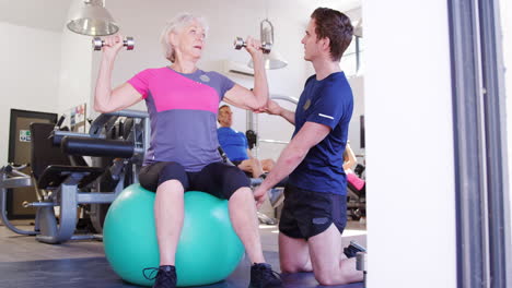 Senior-Woman-Exercising-On-Swiss-Ball-With-Weights-Being-Encouraged-By-Personal-Trainer-In-Gym