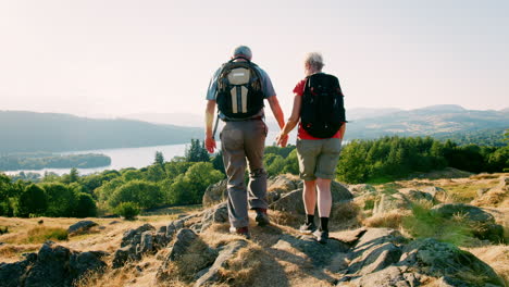 Slow-Motion-Rear-View-Shot-Of-Senior-Couple-Standing-At-Top-Of-Hill-Looking-At-Beautiful-Countryside-On-Hike-Through-Lake-District-In-UK