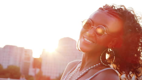 Young-black-woman-wearing-sunglasses-looking-to-camera-laughing-on-a-London-street-at-the-golden-hour,-close-up