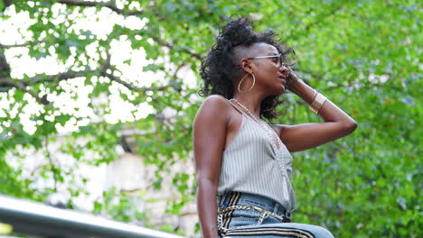 Trendy-young-black-woman-wearing-camisole,-side-stripe-jeans-and-sunglasses,-sitting-on-handrail-playing-with-her-hair,-low-angle