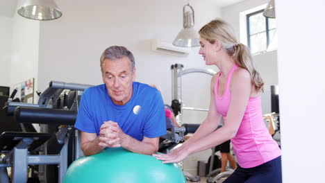 Senior-Man-Exercising-On-Swiss-Ball-Being-Encouraged-By-Personal-Trainer-In-Gym