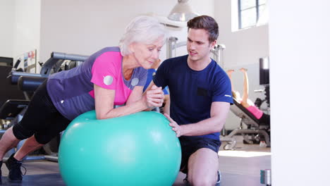 Ältere-Frau-Trainiert-Auf-Einem-Schweizer-Ball-Und-Wird-Von-Einem-Personal-Trainer-Im-Fitnessstudio-Ermutigt
