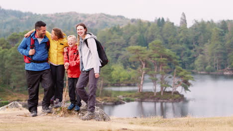 Young-family-on-the-left-of-shot-standing-on-a-rock-by-a-lake-in-the-countryside-looking-to-camera-laughing,-Lake-District,-UK