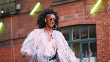 Fashionable-young-black-woman-wearing-multicoloured-fringed-jacket,-sunglasses-and-jeans-turning-around-laughing,-low-angle