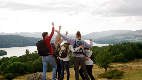 A-multi-ethnic-group-of-five-happy-young-adult-friends-cheer-with-arms-in-the-air,-high-five-and-embrace-at-the-summit-during-a-mountain-hike
