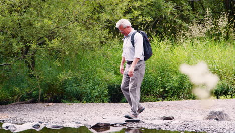 Slow-Motion-Shot-Of-Senior-Man-Using-Stepping-Stones-To-Cross-River-Whilst-Hiking-In-UK-Lake-District