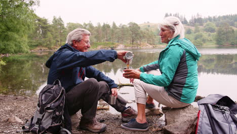 Senior-couple-sitting-by-a-lake-drinking-coffee-during-camping-holiday,-Lake-District,-UK