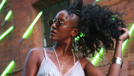 Fashionable-young-black-woman-wearing-round-sunglasses-playing-with-her-hair,-smiling-and-lifting-sunglasses,-low-angle,-close-up,-blurred-neon-strip-lights-in-background