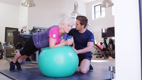 Ältere-Frau-Trainiert-Auf-Einem-Schweizer-Ball-Und-Wird-Von-Einem-Personal-Trainer-Im-Fitnessstudio-Ermutigt