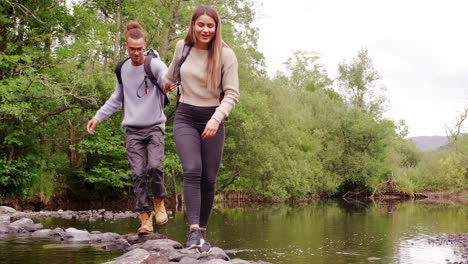 A-young-adult-couple-hold-hands-while-crossing-a-stream-balancing-on-stones-during-a-hike