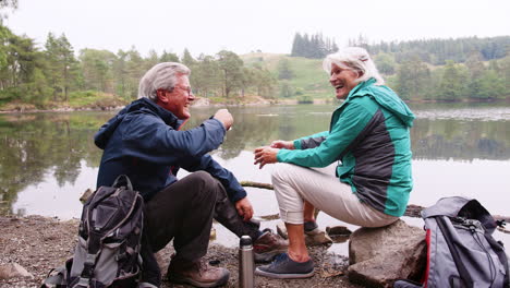 Senior-couple-sitting-on-rocks-by-a-lake-laughing-and-talking-during-a-camping-holiday,-Lake-District,-UK
