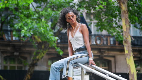 Trendy-young-black-woman-wearing-camisole-and-side-stripe-jeans,-sitting-outdoors-on-handrail