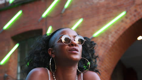 Fashionable-young-black-woman-wearing-round-sunglasses-sitting-on-the-street,-low-angle,-close-up,-blurred-neon-strip-lights-in-background