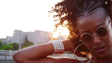 Close-up-of-young-black-woman-wearing-sunglasses-plying-with-her-hair,-looking-to-camera-on-a-London-street-at-the-golden-hour