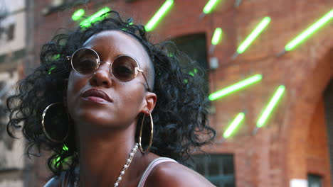 Fashionable-young-black-woman-wearing-round-sunglasses-posing-to-camera-in-an-urban-street,-close-up,-low-angle,-blurred-neon-strip-lights-in-background