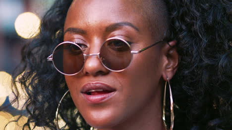 Young-black-woman-wearing-round-sunglasses,-turning-around-and-laughing,-close-up-head-shot,-bokeh-lights-in-background