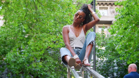 Trendy-young-black-woman-wearing-camisole-and-side-stripe-jeans,-sitting-on-handrail-in-green-city-street