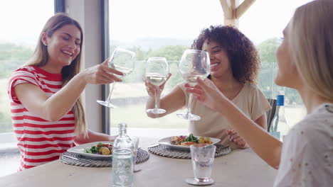 Three-happy-young-adult-women-making-a-toast,-talking-and-eating-at-a-dinner-party