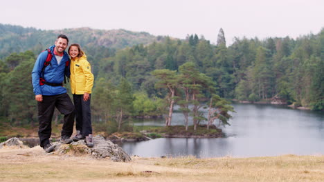 Caucasian-father-and-daughter-on-the-left-of-shot-standing-on-rock-by-a-lake-in-the-countryside-looking-to-camera,-Lake-District,-UK