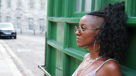 Trendy-young-black-woman-wearing-round-sunglasses-and-camisole-leaning-on-green-wall-in-a-city-street,-side-view,-head-and-shoulders