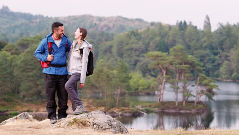 Young-adult-couple-standing-on-the-left-of-shot-against-a-lake-view,-laughing-to-camera,-Lake-District,-UK