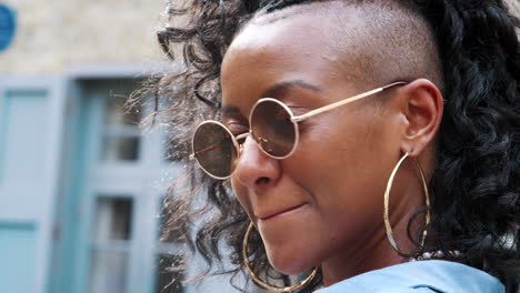 Trendy-young-black-woman-wearing-round-sunglasses-turning-to-look-back-to-the-camera,-head-and-shoulders,-close-up