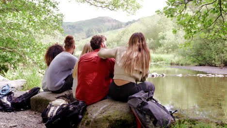Five-young-adult-friends-admire-the-landscape-while-taking-a-break-sitting-on-rocks-by-a-river-during-a-hike,-back-view