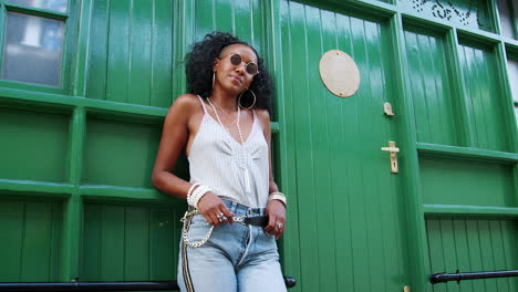 Fashionable-young-black-woman-wearing-sunglasses-leaning-by-green-door-outdoors-lifts-sunglasses,-low-angle