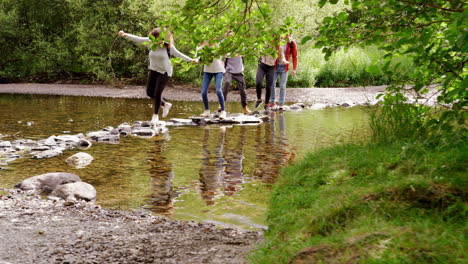 Five-young-adult-friends-hold-hands-and-help-each-other-cross-a-stream-balancing-on-stones-during-a-hike
