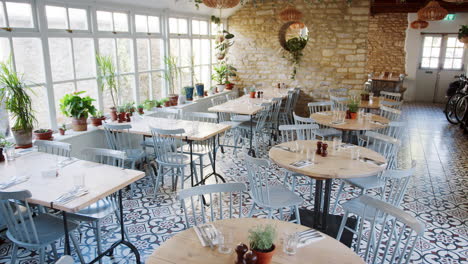 Runde-Tische-Und-Blaue-Stühle-In-Einem-Leeren-Restaurant-Mit-Gemusterten-Bodenfliesen-Und-Zimmerpflanzen,-Die-Auf-Der-Fensterbank-Wachsen