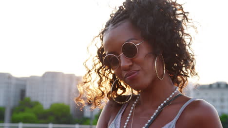 Young-black-woman-wearing-sunglasses-looking-to-camera-and-smiling-on-a-London-street-at-sunset,-close-up