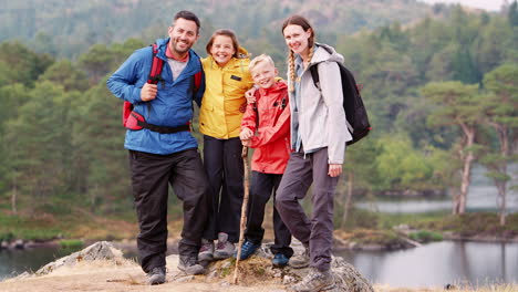 Young-family-standing-on-a-rock-by-a-lake-in-the-countryside-looking-to-camera-laughing,-Lake-District,-UK