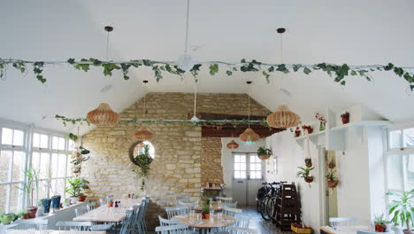 Empty-tables-and-chairs-in-a-restaurant-dining-space-in-daylight,-with-feature-stone-walls-and-tiled-floor,-tilt-shot