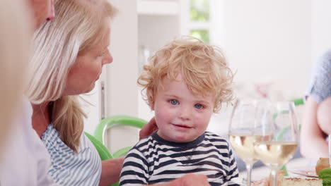 Grandmother-Playing-With-Grandson-As-They-Sit-At-Table-For-Family-Meal