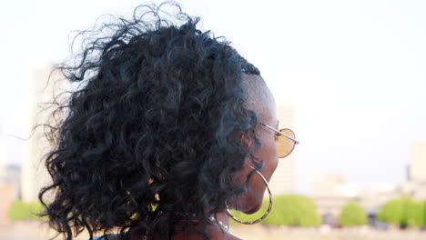 Trendy-young-black-woman-in-camisole-and-sunglasses-walking-over-bridge,-looking-back-and-talking-to-camera,-close-up,-follow-shot