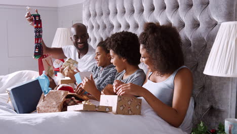 Young-mixed-race-family-sitting-up-in-bed-together-unwrapping-presents-on-Christmas-morning,-side-view,-close-up