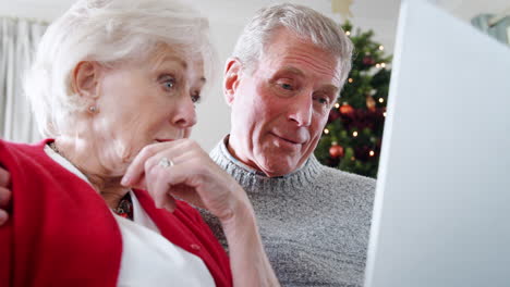 Nahaufnahme-Eines-älteren-Paares,-Das-Auf-Dem-Sofa-Sitzt-Und-Mit-Einem-Laptop-Zu-Hause-Waren-Oder-Dienstleistungen-Kauft,-Mit-Einem-Weihnachtsbaum-Im-Hintergrund