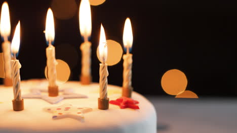 Nahaufnahme-Von-Brennenden-Kerzen-Auf-Einem-Weißen,-Dekorierten-Geburtstagskuchen,-Bokeh-Lichter-Im-Hintergrund