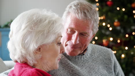 Close-Up-Of-Senior-Couple-Sitting-On-Sofa-At-Home-Chatting-With-Christmas-Tree-In-Background