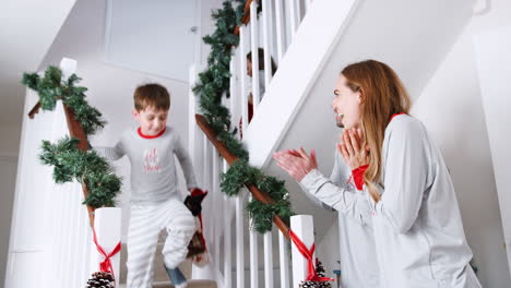 Padres-Saludando-A-Niños-Emocionados-En-Pijama-Corriendo-Escaleras-Abajo-Sosteniendo-Medias-En-La-Mañana-De-Navidad